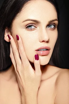 Close-up van het gezicht van een vrouw met een lichte huid, blauwe ogen en donker haar. Ze raakt haar wang aan met haar hand, waarop maroon nagellak te zien is. De uitdrukking is contemplatief.