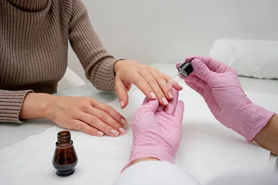 Een persoon in een bruine trui krijgt een manicure. Een andere persoon met roze handschoenen brengt met een pipet nagelriemolie aan. De sfeer is kalm en professioneel.